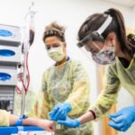 Two students wearing facemasks, one clear plastic, the other a surgical mask, and wearing protective plastic robes and gloves prepare to draw blood in phlebotomy class.