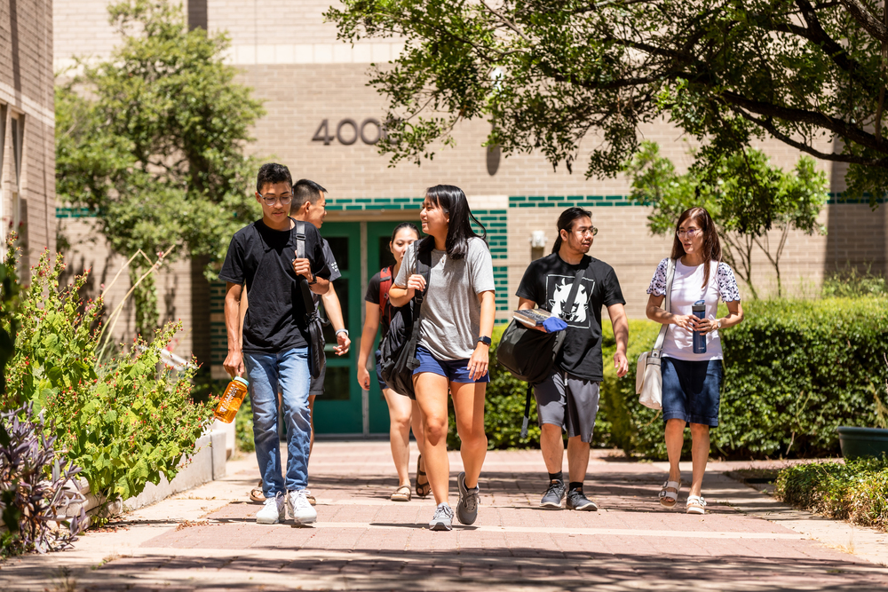Several men and women students walk along a walkway outside a tan brick building on the Austin Community College Northridge Campus.