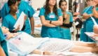 A class of student nurses in teal scrubs follow a professor's lecture. They surround a dummy patient in a hospital gown in a training facility.