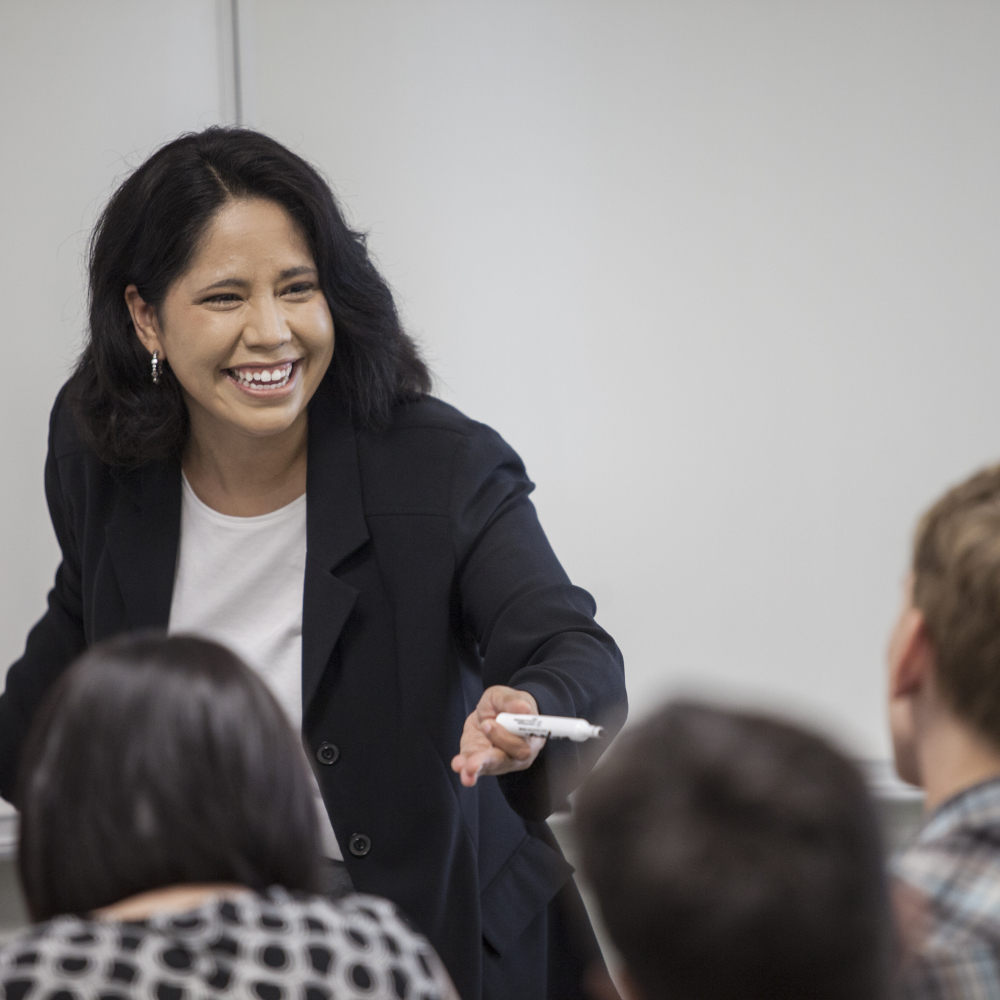 A teacher with dark hair, wearing a black blazer and white shirt, smiles as she talks with students in her classroom at Austin Community College.
