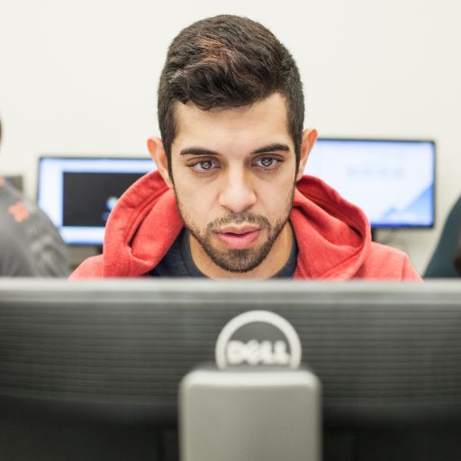 a young male student wearing a red hoodie looks intently into a computer monitor.