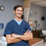 a male nursing student wearing dark blue scrubs poses in a hospital simulation classroom.