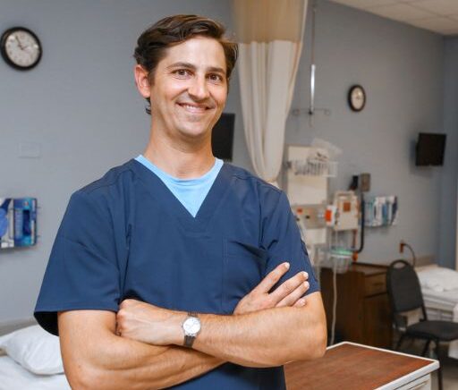 a male nursing student wearing dark blue scrubs poses in a hospital simulation classroom.