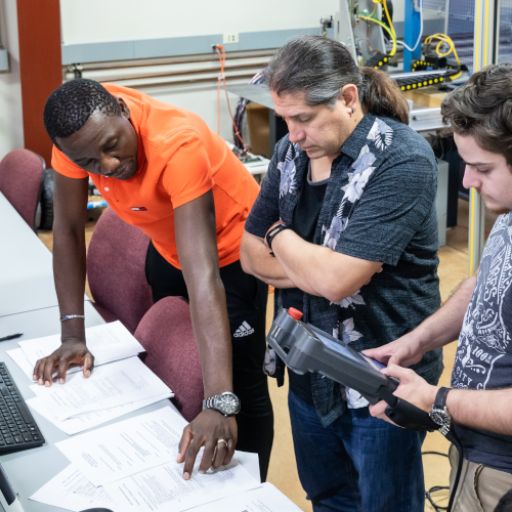 A Black student in an orange shirt, a white student in a band t-shirt holding a handheld monitoring device, and a professor in a black and white Hawaiian shirt look over paperwork in a manufacturing classroom.