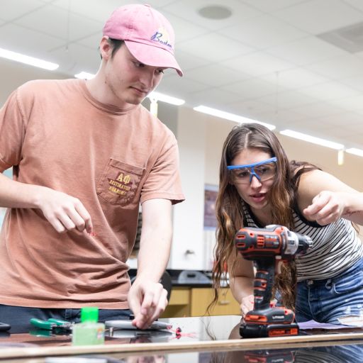 A male student in a pink ball cap and a female student with safety goggles and a striped shirt participate in an ACC Engineering physics lab project.