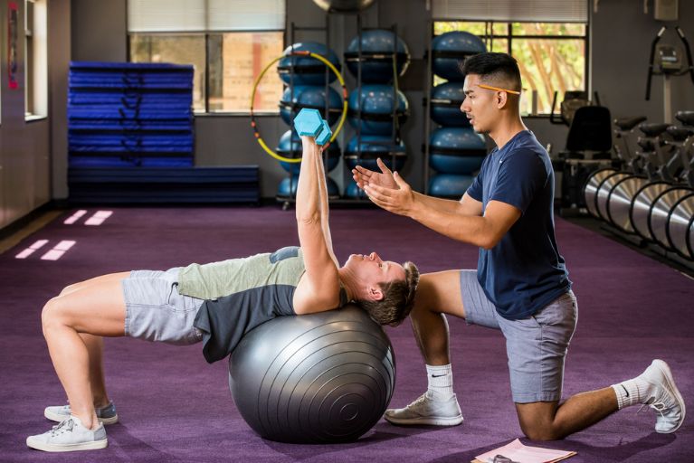 A male personal trainer wearing a blue shirt and gray shorts supports a female client as she does arm weights while balanced on her back on an exercise ball.