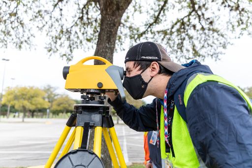 A male student wearing a yellow safety vest over a blue coat, and a backward baseball cap peers through a surveying tool in a GIS lab.