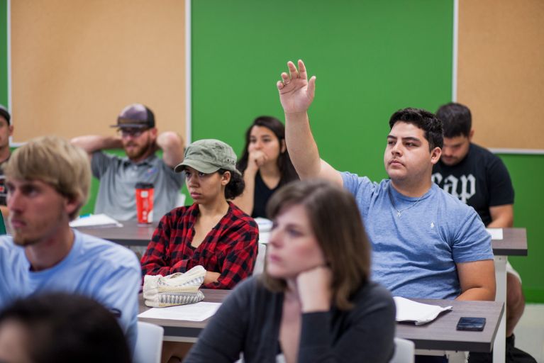 A male student in a blue shirt raises his hand in a government classroom of diverse students at Austin Community College
