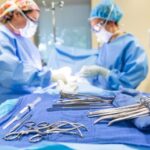 Two students in blue scrubs, face masks, and head coverings practice with medical instruments, including scalpels and other devices in a surgical technology class at ACC.