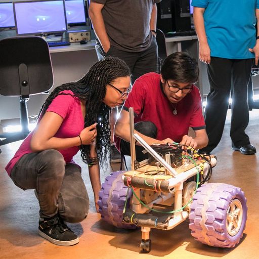 A female student and a male student in ACC's engineering technology program kneel down to operate a robot with giant knobby wheels while two onlookers stand by.