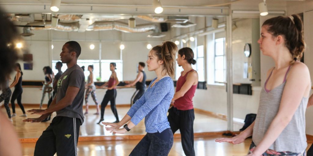 Student dancers, including men and women, practice in a studio with a mirror.