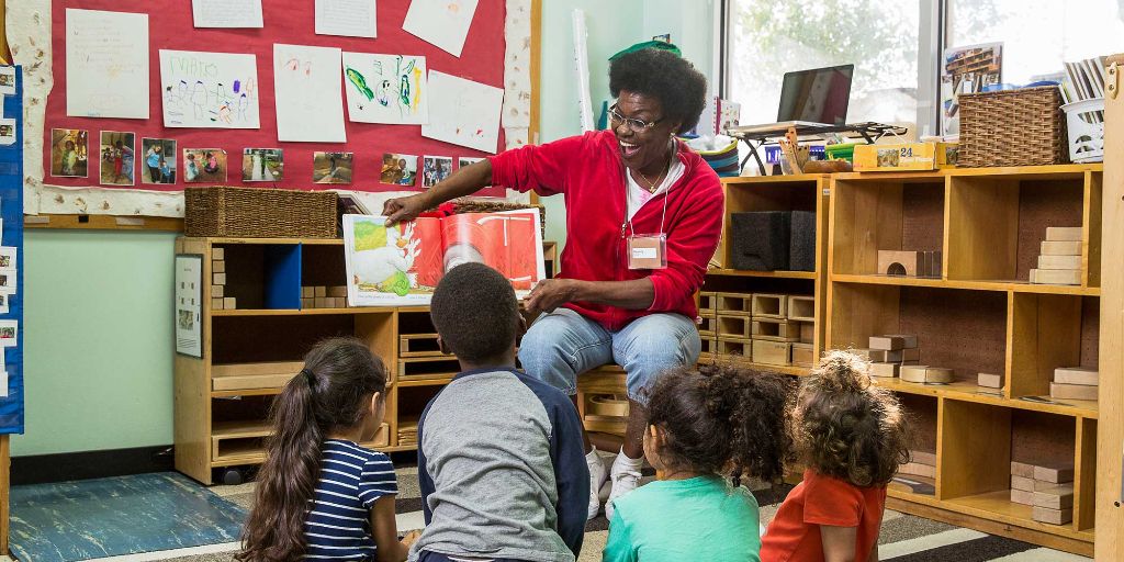 A female teacher in a red pullover and jeans reads a picture book to preschoolers in a daycare classroom filed with blocks, toys, and books.