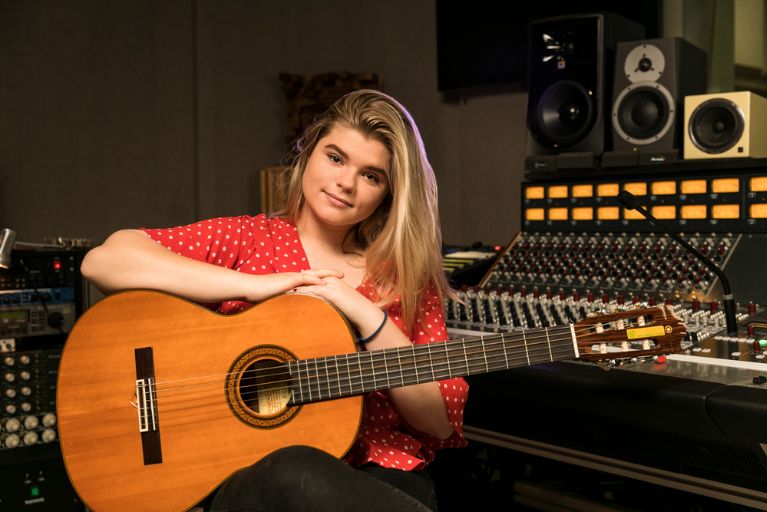 A female student with long blond hair, red blouse, and acoustic guitar sits in front of an audio recording control board.