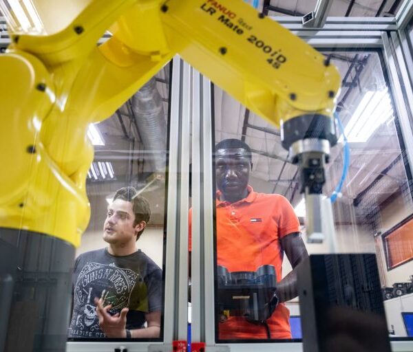 Two male students, one wearing a Guns n Roses t-shirt and another in an orange polo, send instructions to a yellow robotic arm in a glass and steel enclosure.