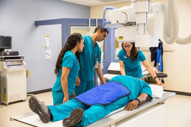 Two female students and one male student in teal scrubs practice imaging a fellow student in teal scrubs with a blue lead-lined apron over him under a diagnostic imaging machine.
