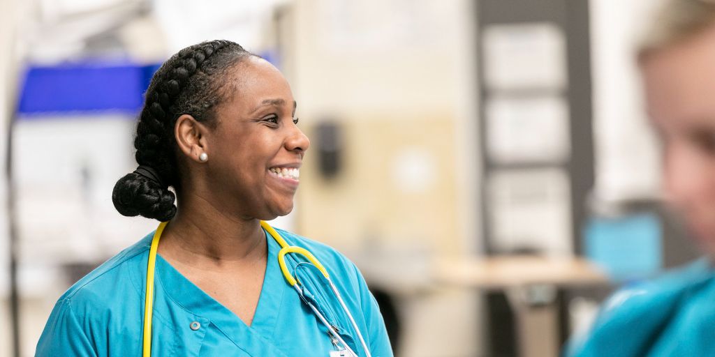A vocational nursing student teal scrubs with a yellow stethoscope smiles with a classmate just out of frame.
