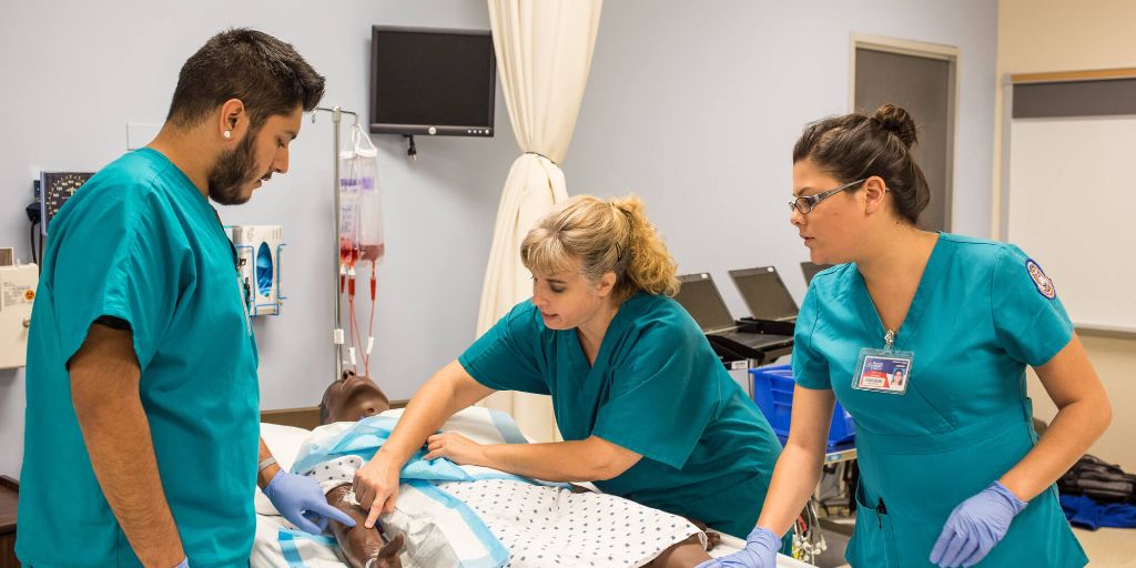 Three nurses in teal scrubs, two women and one man, learn how to check vital signs on a mannequin in a hospital bed training lab.