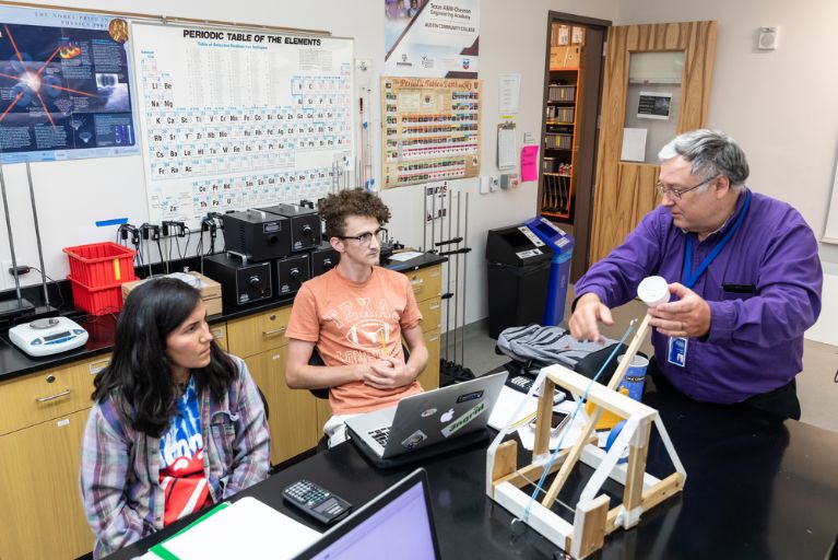 A physics professor in a purple shirt speaks with physics students, one a woman, one a man, in a phyics lab at the ACC Rio Grande campus.