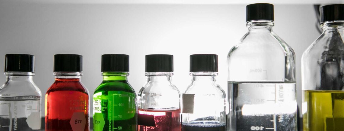 Different colored chemicals sit in clear bottles on a shelf.