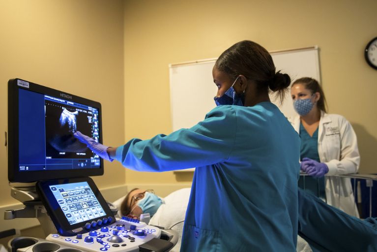 A sonographer in teal scrubs gestures at a sonography image as she scans a patient, while a nurse looks on in the background.