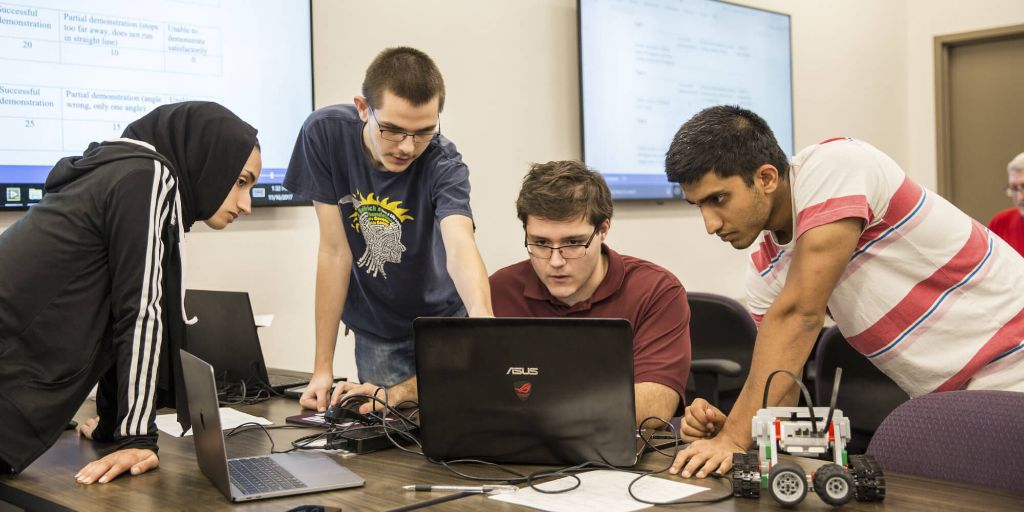 Four students, three young men and one young woman, pore over a laptop and a remote-control toy vehicle in an engineering class.