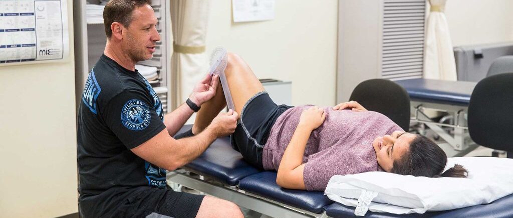 A physical therapy assistant student practices on a volunteer student lying on a raised gurney.