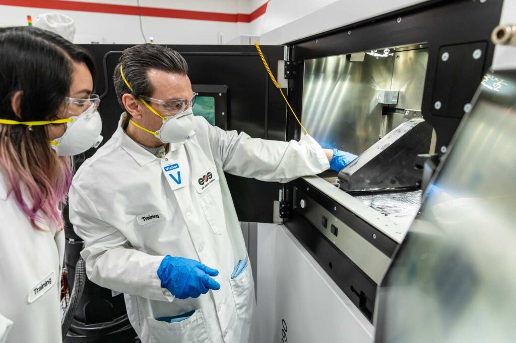 A man and woman in labcoats and N95 masks look at 3D printing equipment in ACC's additive manufacturing program. 