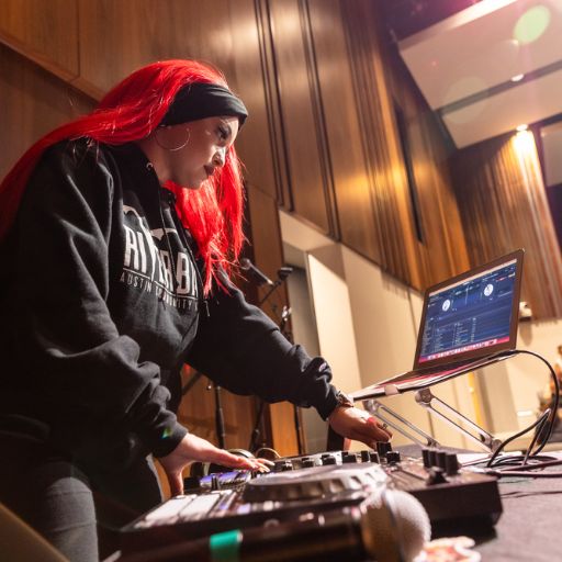 an ACC student with long bright red hair operates a turntable and monitor as she djs a hip-hop celebration in the Audio Technology and Industry program.