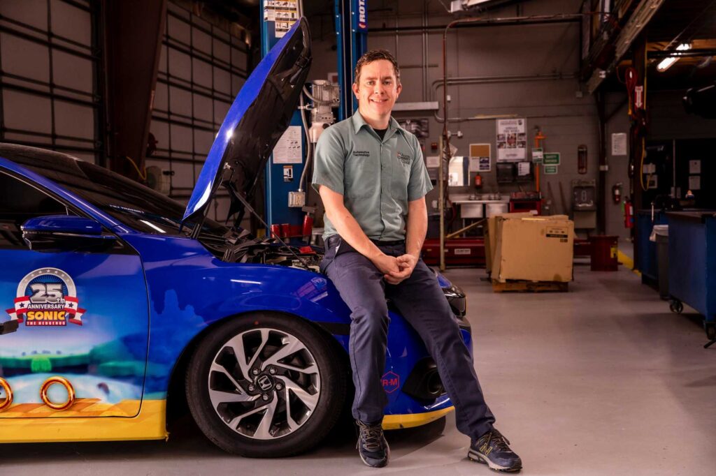 Honda PACT institute instructor sits on the front fender of a blue car with the hood up.