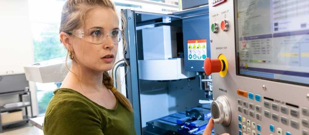 A female student in a green t shirt and safety glasses operates a manufacturing machine.