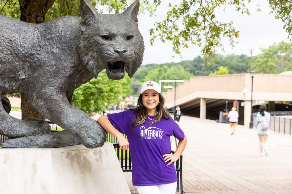 An ACC transfer student poses on the Texas State campus in front of a statue of the Bobcat mascot.