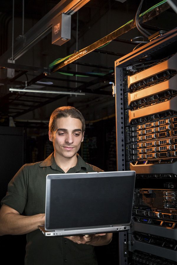 A male student works on a laptop in a network server room.