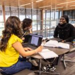 A female student with brown hair and a yellow shirt sits in the foreground, a male student to her left, and facing an African American female student as they study at a table overlooking windows.