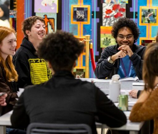 smiling students sit around a table in front of a colorful mural discussing the topics in the great questions seminar.