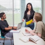 Three business students work on a project together with a laptop and notebooks in a classroom with a white table and wide windows