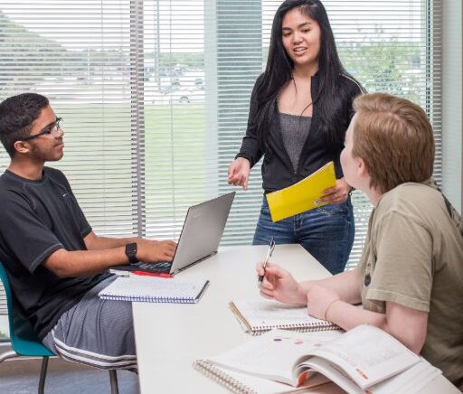 Three business students work on a project together with a laptop and notebooks in a classroom with a white table and wide windows