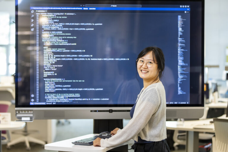 Female Asian student poses in front of large monitor showing lines of code.