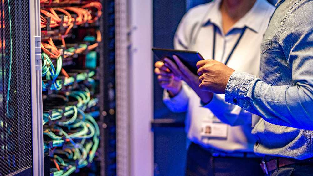 Two men stand in front of a server rack, one holding a tablet while discussing technology.