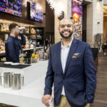 A smiling man in a blue blazer, khakis, and glasses poses in front of a hotel bar.