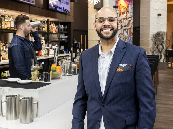 A smiling man in a blue blazer, khakis, and glasses poses in front of a hotel bar.