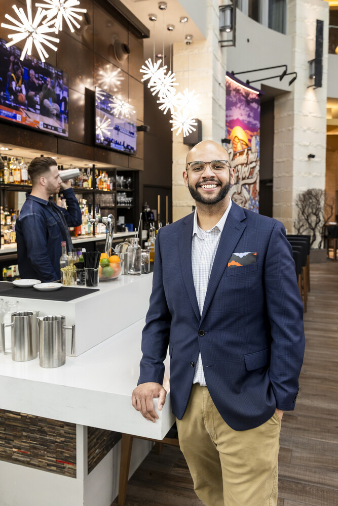 Julian Richmond, ACC hospitality management graduate, wears a blue work blazer, khakis, and glasses as he poses in front of a hotel bar.
