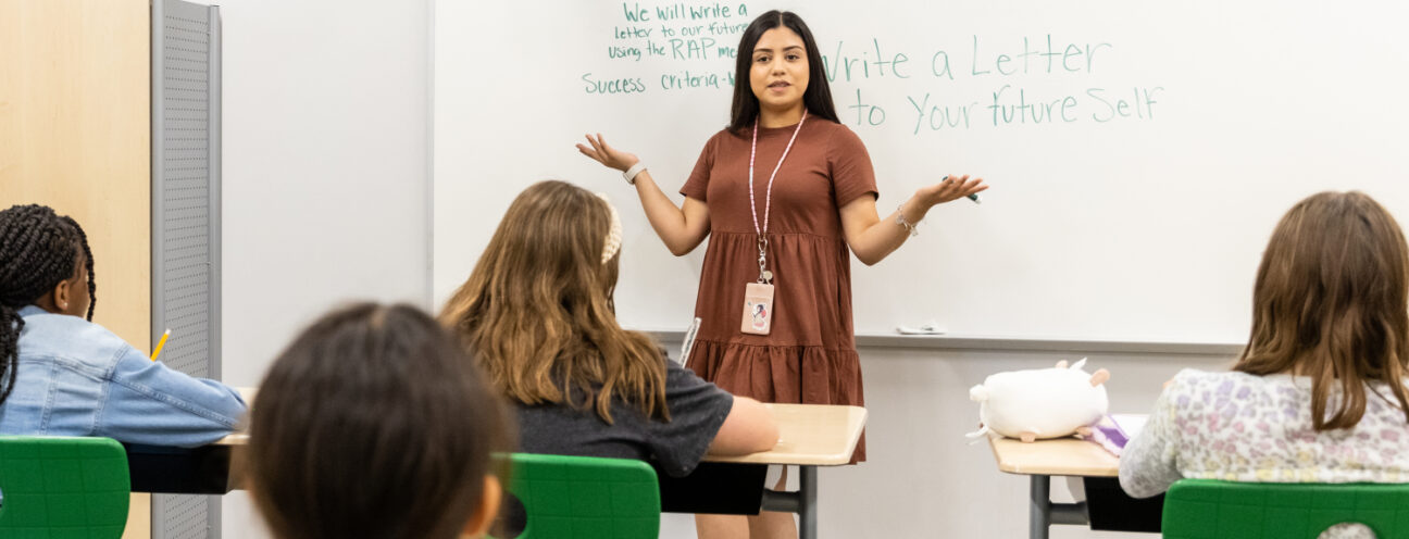 A female student teacher with brown hair and a brown dress stands in front of a classroom of fourth and fifth graders working on a lesson on writing a letter to your future self; she is learning classroom management skills