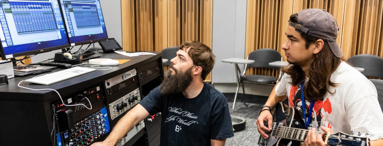 A man with long hair and a baseball cap backwards plays guitar as another man with a long beard manages a soundboard in a studio.