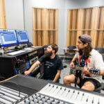 A man with long hair and a baseball cap backwards plays guitar as another man with a long beard manages a soundboard in a studio.