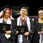 A group of five dual credit graduates stands together in a large indoor arena, smiling and holding their diplomas. They are dressed in black graduation gowns with white stoles saying Austin Community College.