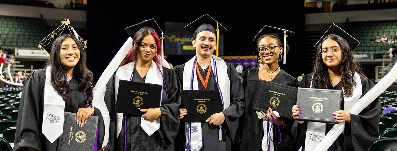 A group of five dual credit graduates stands together in a large indoor arena, smiling and holding their diplomas. They are dressed in black graduation gowns with white stoles saying Austin Community College.