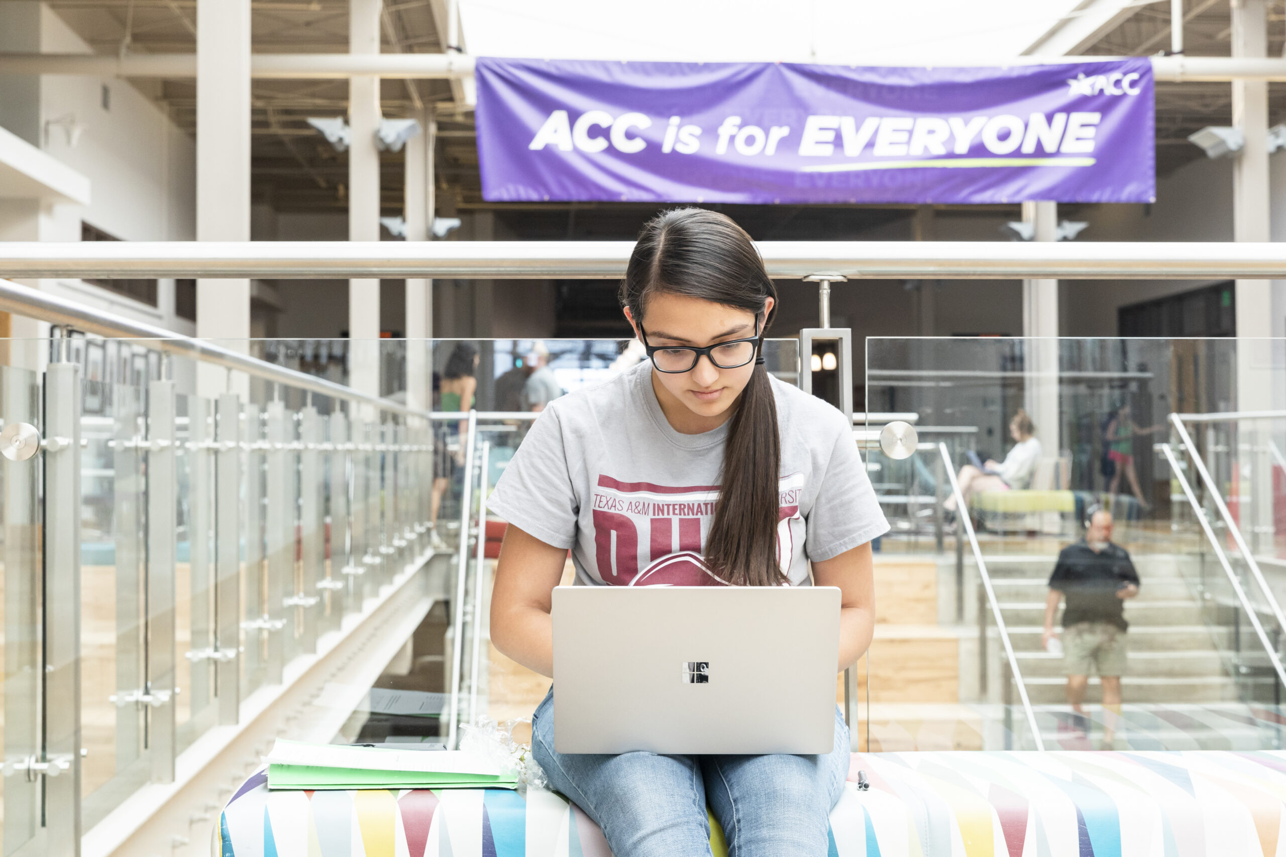 Female student with long brown hair and glasses, wearing a gray Texas A&M T shirt sits on a brightly colored bench in front of wooden stairs and a purple banner that says ACC is for Everyone, working on her laptop.