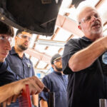 Three automotive technology degree students wearing blue uniform shirts and protective goggles watch an instructor work on an engine.