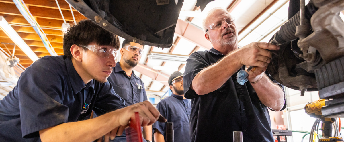Three automotive technology degree students wearing blue uniform shirts and protective goggles watch an instructor work on an engine.